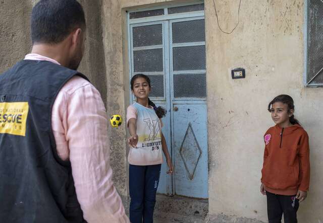Ein Mann mit einer Weste spricht vor einem Gebäude mit zwei Kindern, die vor einer blauen Tür stehen.